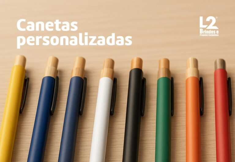 Uma fileira de canetas personalizadas de diversas cores (amarelo, azul, branco, preto, verde, laranja, vermelho) com detalhes em bambu, dispostas sobre uma superfície de madeira, com o texto Canetas personalizadas e o logo L2 Brindes.