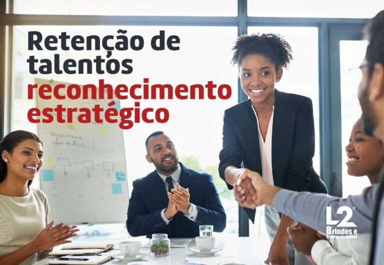Uma mulher sorridente, vestindo um blazer preto, aperta a mão de um colega em uma mesa de reunião, enquanto outros colegas, incluindo um homem aplaudindo, observam. A imagem tem sobreposta a frase Retenção de talentos reconhecimento estratégico em destaque, e o logo L2 Brindes e Presentes Exclusivos no canto inferior direito.