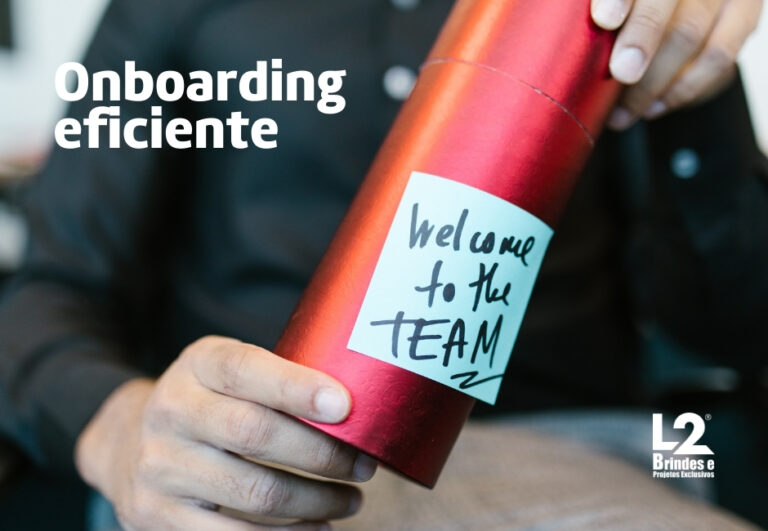 Mãos segurando um tubo vermelho brilhante com um adesivo azul claro que diz "Welcome to the TEAM" em caligrafia preta. No canto superior esquerdo, o texto "Onboarding eficiente" em branco. No canto inferior direito, um logotipo "L2 Brindes e Projetos Exclusivos".